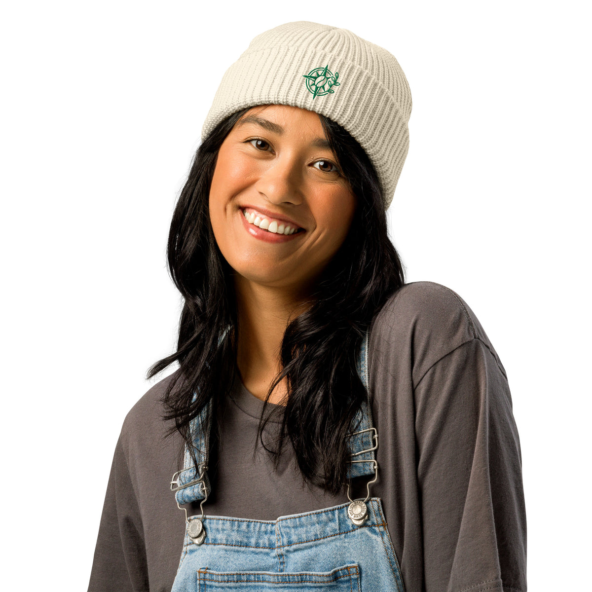 Woman wearing a beige knit beanie with a logo, gray shirt, and blue denim overalls on a white background