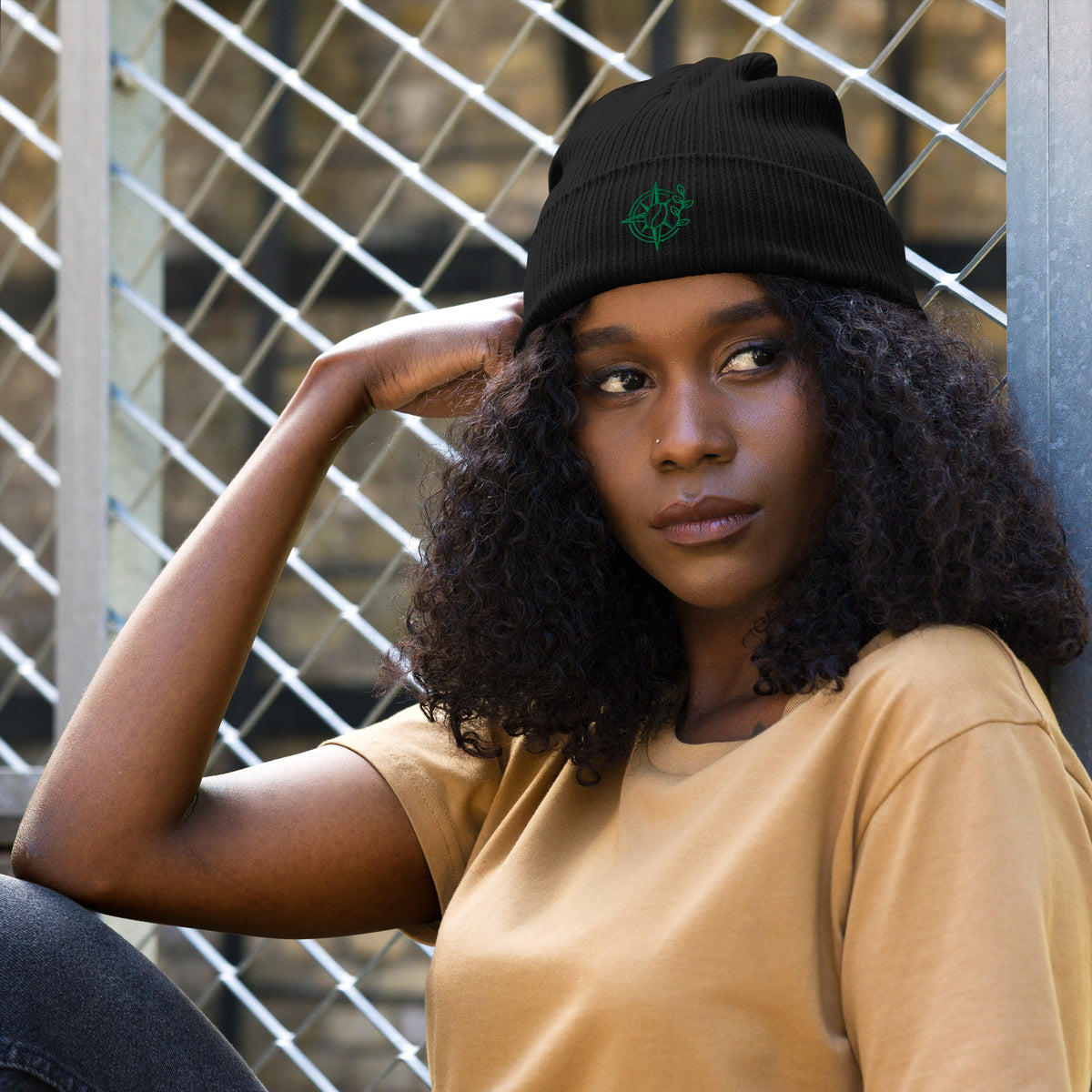 Woman wearing a black beanie with 'wild fire.' text and a mustard yellow shirt against a metal fence background.