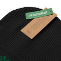 Close-up of a black ribbed knit beanie with a green 'I'M SUSTAINABLE' hangtag and a brown paper tag reading 'ORGANIC COTTONS' on a white background.