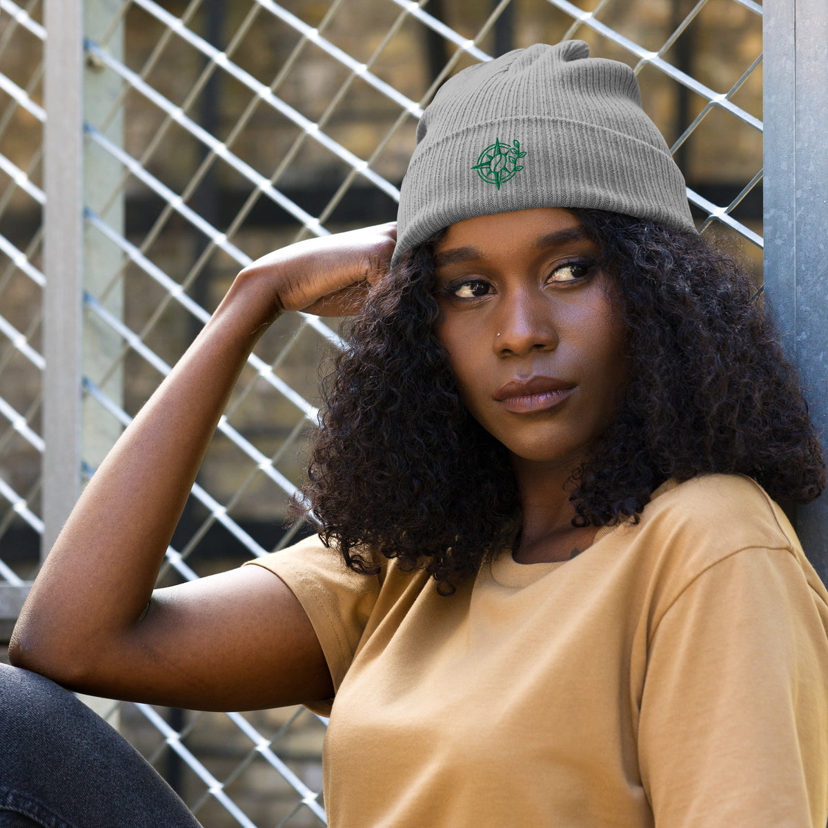 Woman wearing a gray beanie with a logo, sitting against a metal fence.
