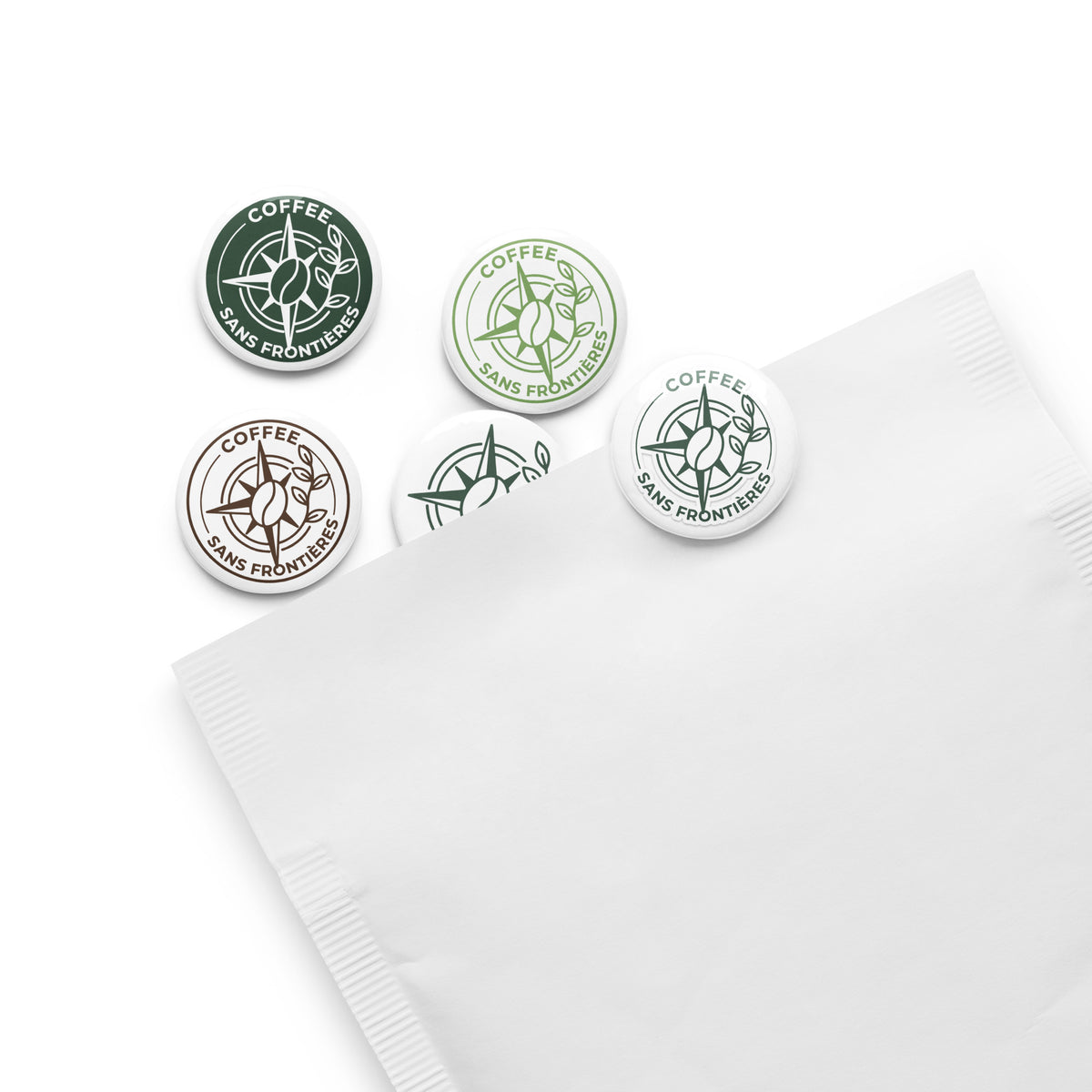 Five round branded buttons or badges with compass-style graphics and the words 'COFFEE SANS FRONTIERS' are arranged on a white surface next to a white envelope or card.