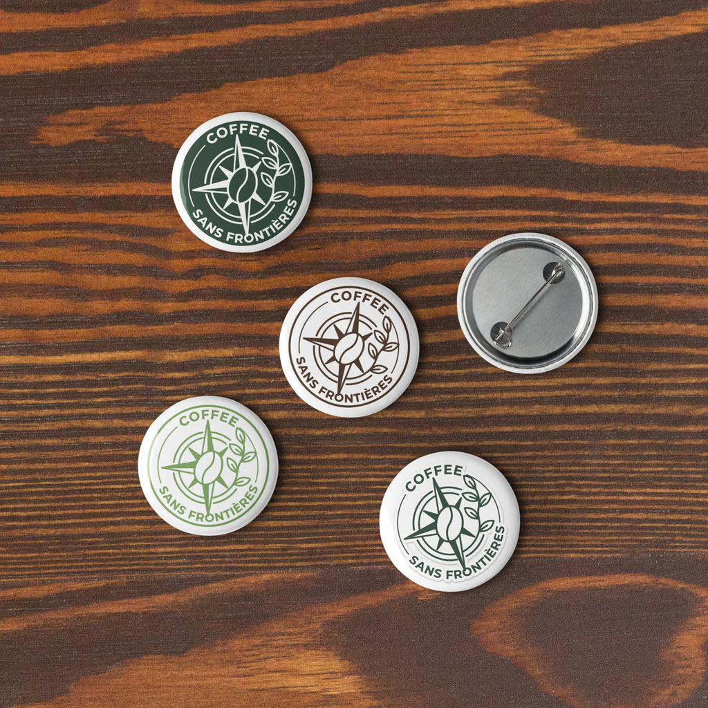 Four round pin-back buttons with the Coffee Sans Frontiers logo are arranged on a dark wood surface. Three buttons show the front design, and one is turned over to show the silver metal pin-back mechanism.