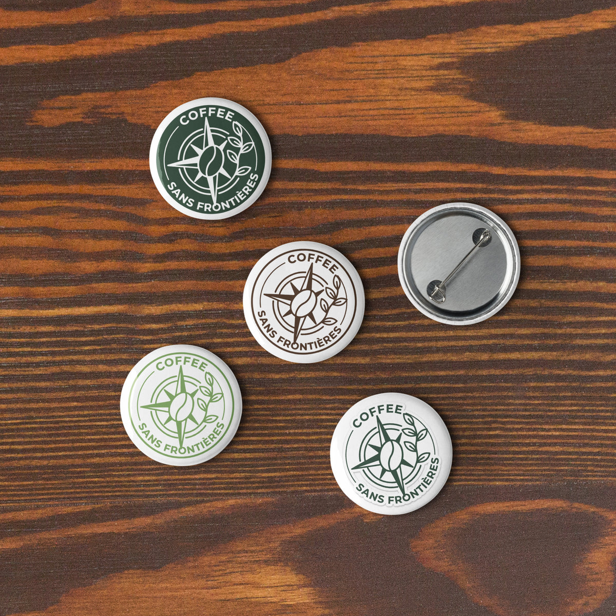 Four round pin-back buttons with the Coffee Sans Frontiers logo are arranged on a dark wood surface. Three buttons show the front design, and one is turned over to show the silver metal pin-back mechanism.