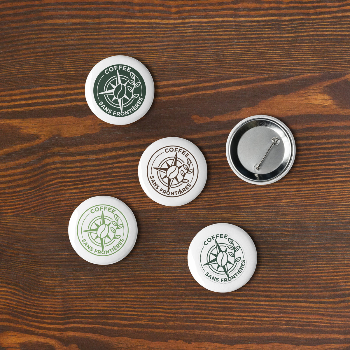 Five round pin-back buttons with the Coffee Sans Frontiers logo are arranged on a dark wood background. Three buttons show the white button face with printed green and black graphics, while one button at the top right shows the metal back with pin fastening.